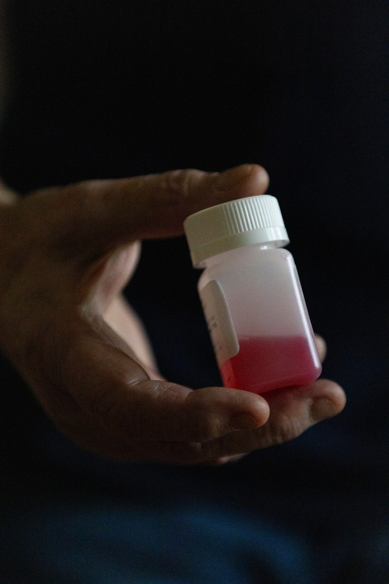 A person's hand holds a small bottle containing a red liquid against a dark background.