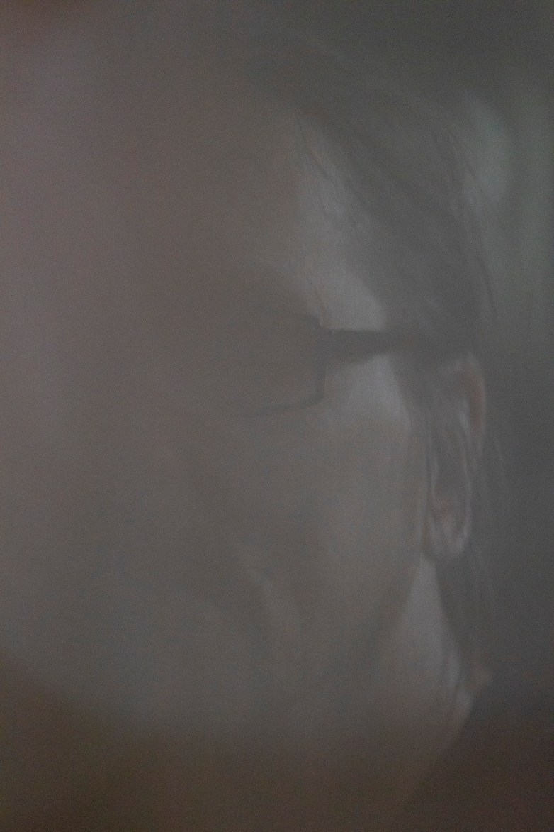 A partially obscured person wearing glasses is seen through a haze, with only part of the face visible.