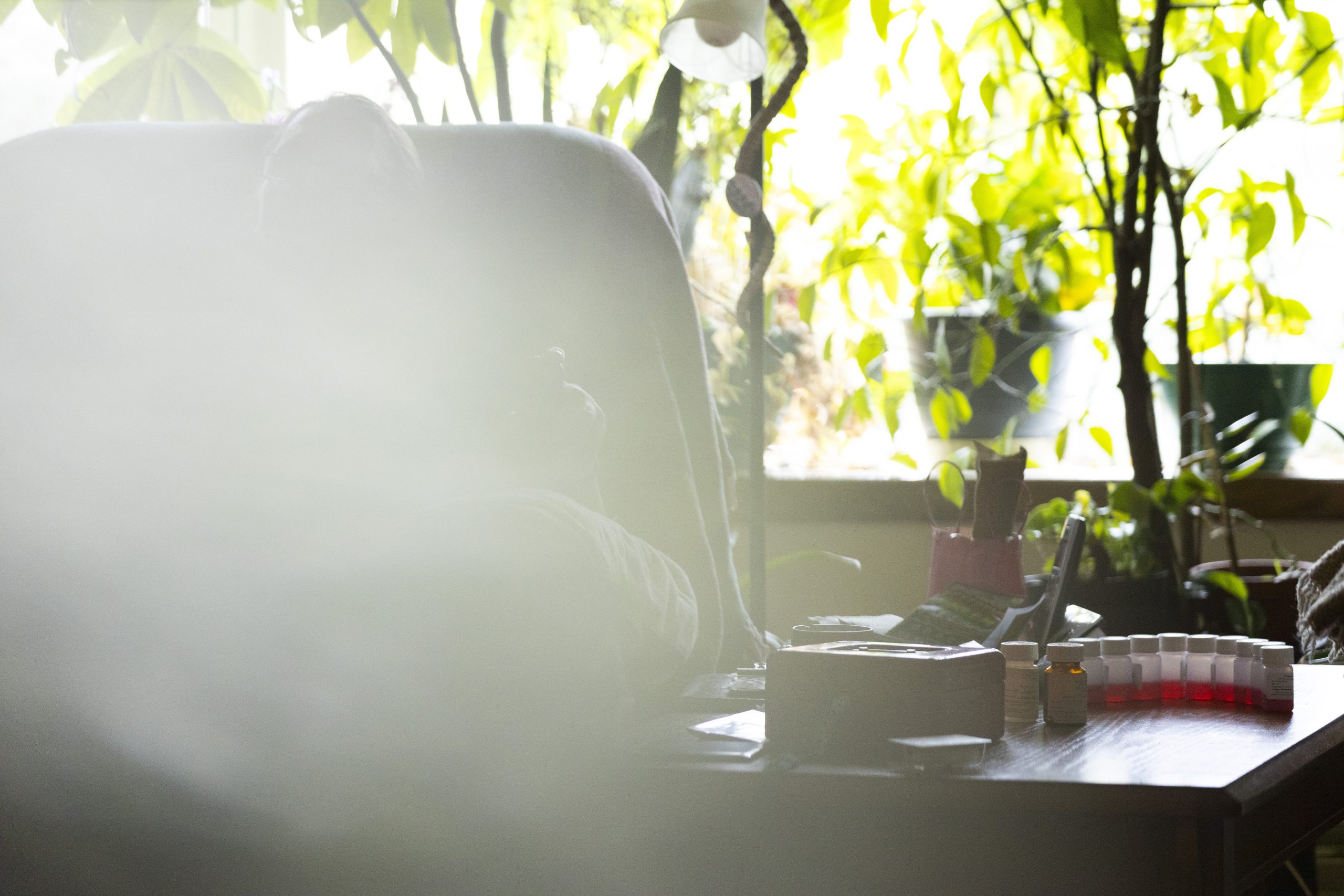 A person obscured by a smoky haze sits in a chair beside a table with multiple small bottles containing red liquid, with houseplants and a window in the background.