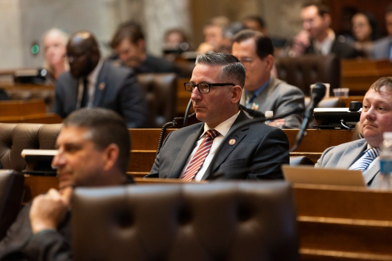 A person wearing glasses and a suit sits among others in a room, with rows of desks and microphones visible.