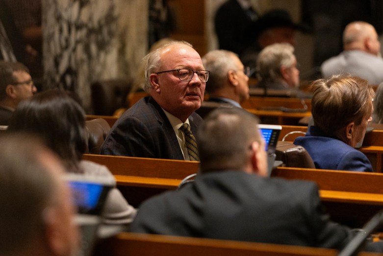 A person wearing glasses and a suit sits among others in rows of desks, with microphones visible.
