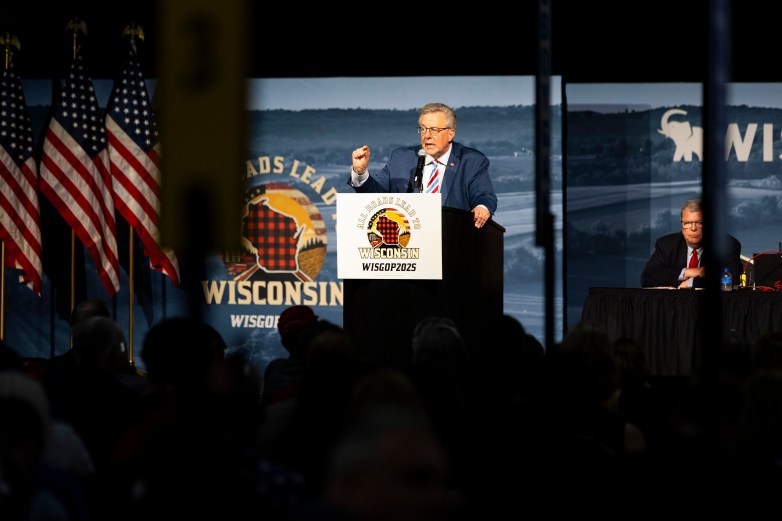A person stands with a hand raised at a podium with a sign reading "All Roads Lead to Wisconsin WISGOP2025" with American flags and an audience visible and another person sitting in the background.