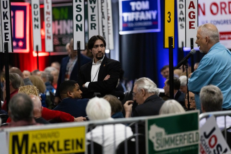 A person sands with arms crossed in the middle of a seated crowd as another person speaks into a microphone at right, with vertical signs and campaign-style posters visible in the background.