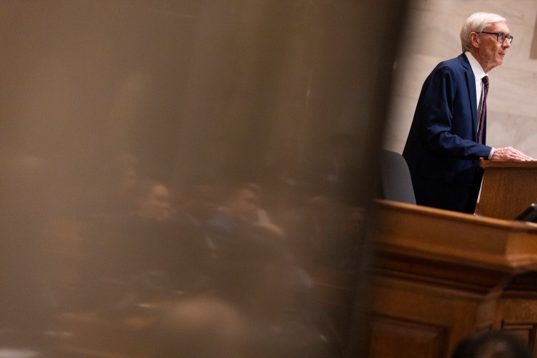 A person in a suit stands at a wooden podium at the right, viewed partially through a blurred foreground with seated people visible.