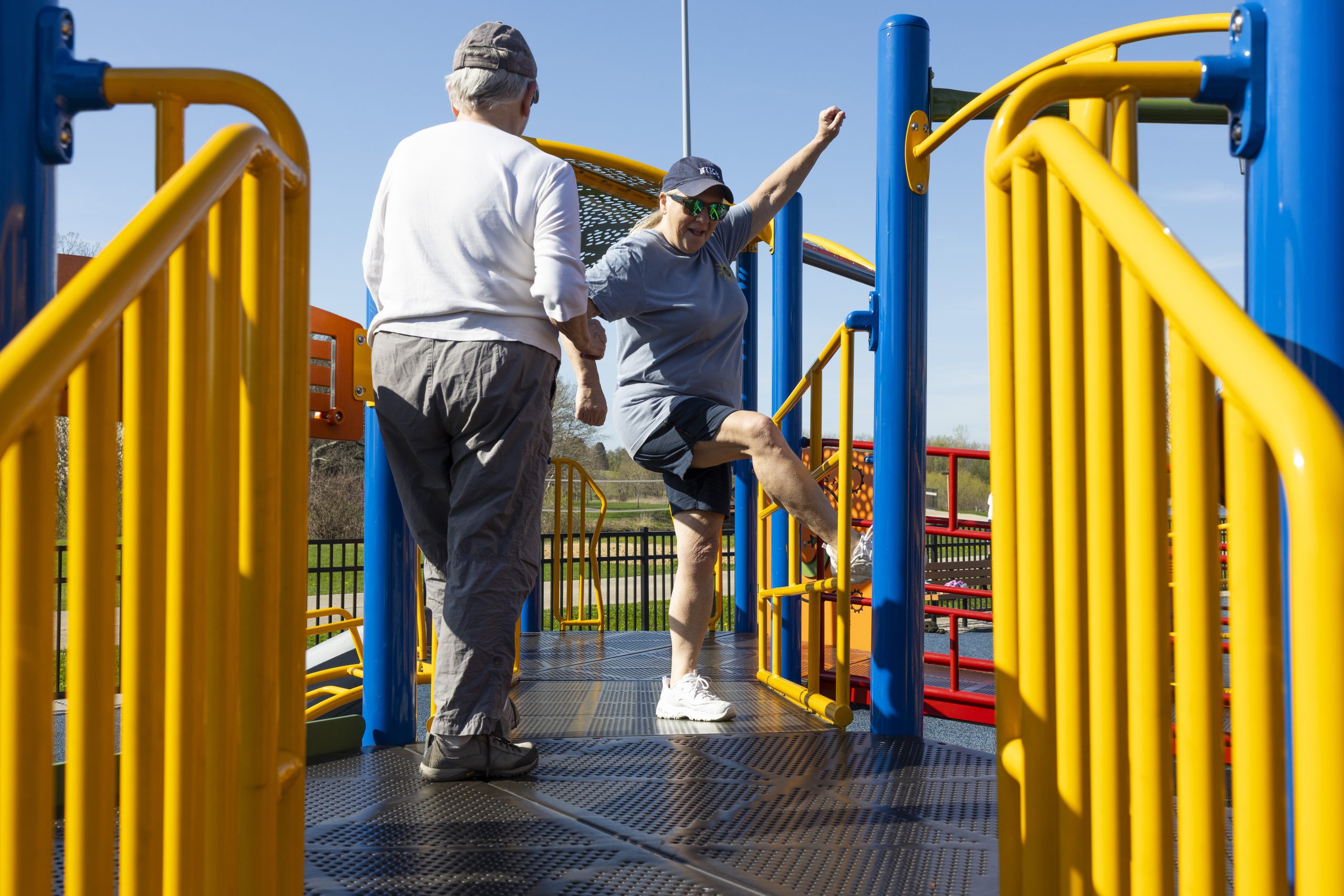 Two people stand on a playground platform with yellow railings and blue poles as one of the people lifts a leg and raises an arm.