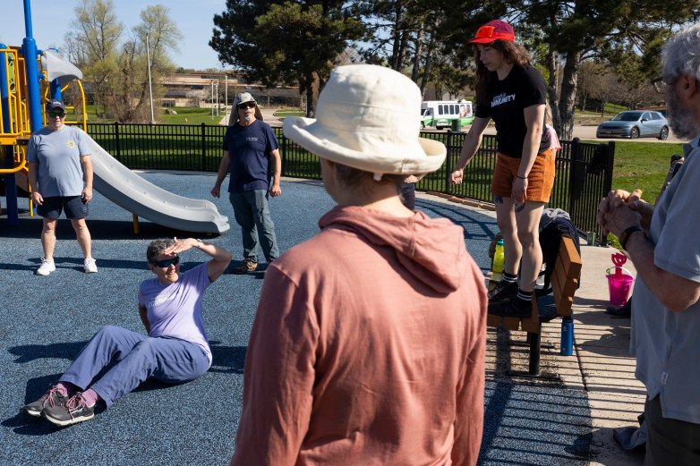 A group of people gather at a playground, with one person sitting on the ground while others stand and watch near a slide and climbing structures.