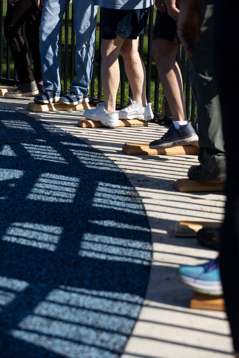 Several people stand on small wooden blocks along a semicircular white and blue surface next to a railing, with shadows cast across the ground.