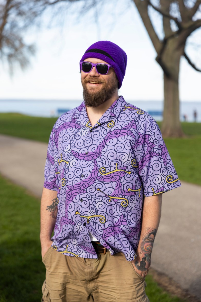 A person wearing a purple hat and shirt and sunglasses stands and smiles near a path near a body of water, with trees and grass in the background.