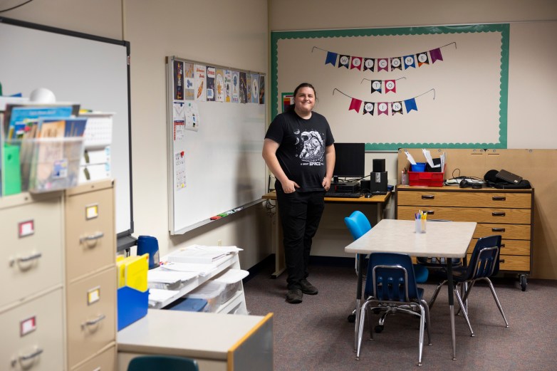 A person wearing a T-shirt with an astronaut graphic stands in a classroom with desks, a whiteboard and a banner reading
