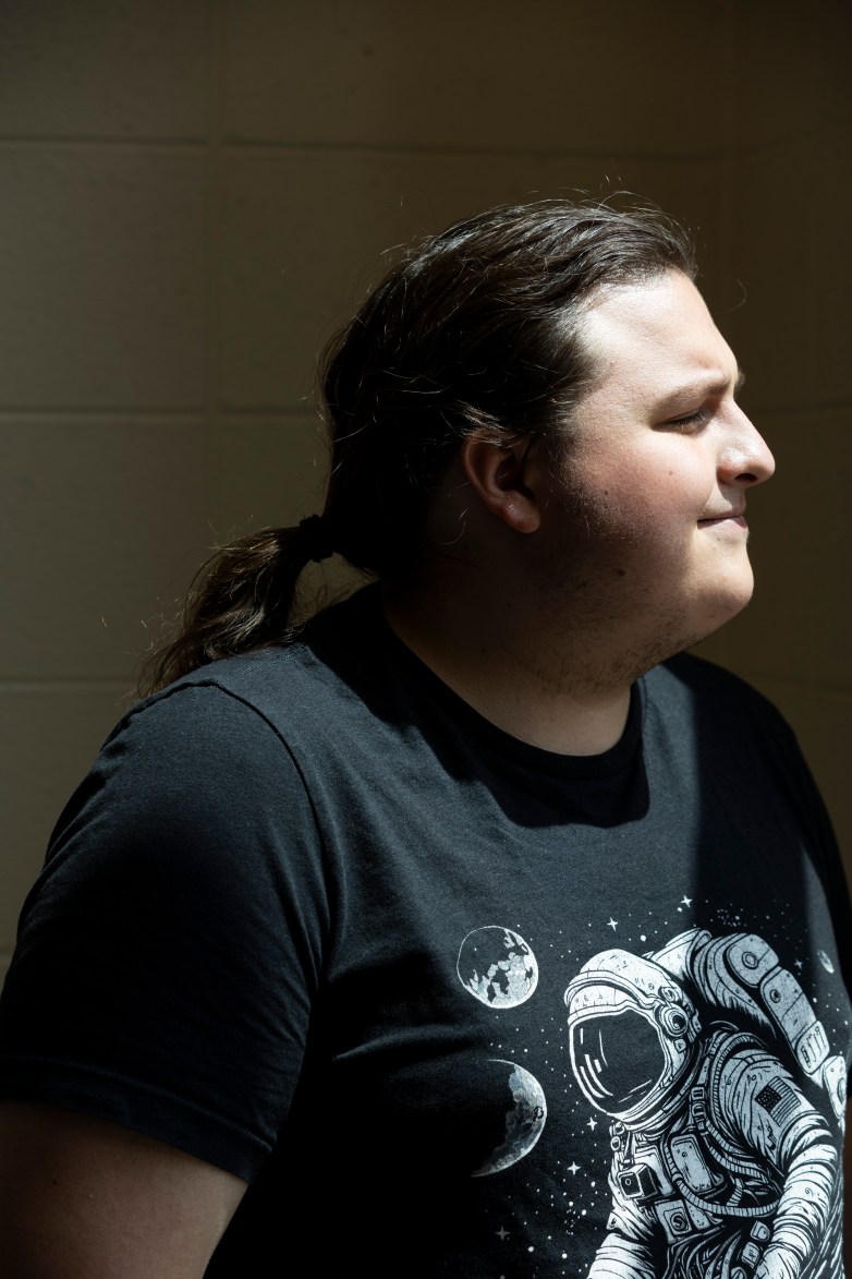 A person with a ponytail wearing a T-shirt with an astronaut graphic stands in sunlight against a tiled wall in profile view.
