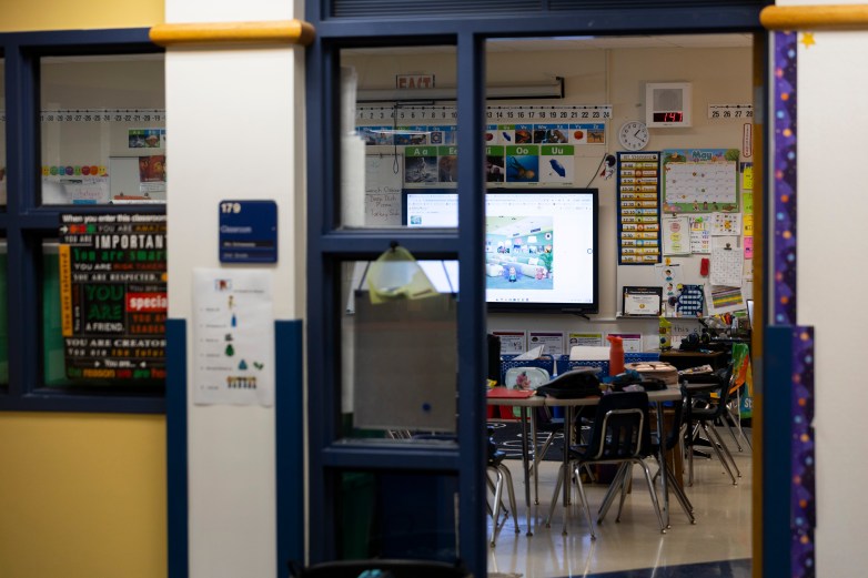An empty classroom with desks, posters and a wall-mounted screen is visible through windows and an open doorway with a sign marked