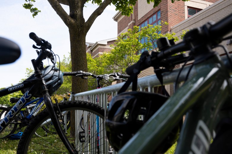 Bicycles and helmets are locked to a metal rack beside trees outside a brick building with large windows.