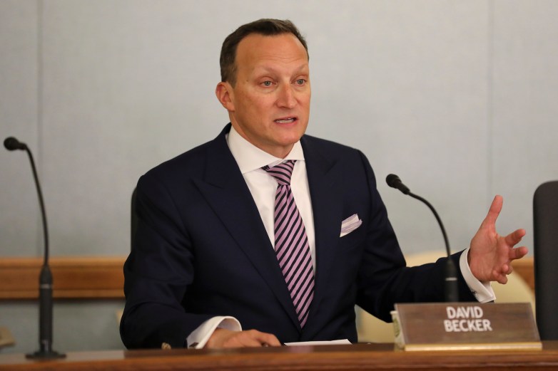 A person in a suit and striped tie sits at a desk between microphones, with a nameplate reading “DAVID BECKER”
