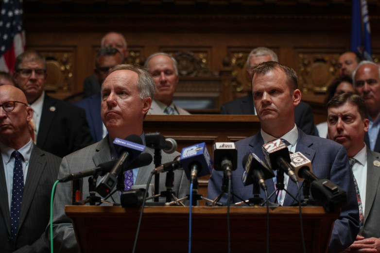 People in suits stand behind a podium with several microphones displaying news station logos inside a wood-paneled room.