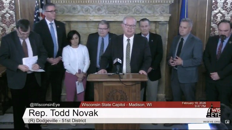 A group of people in suits stand, with one behind a podium with a microphone, with on-screen text reading 
