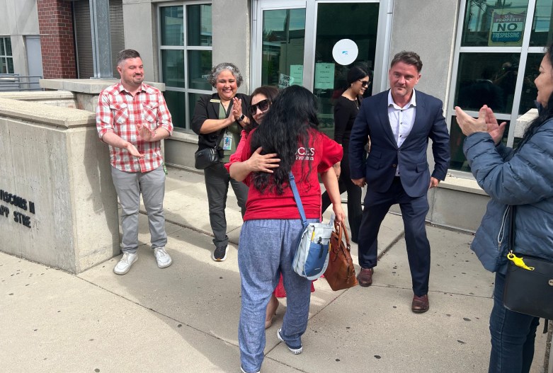 People stand outside a building entrance as one person embraces another; several others clap, and a person holds a brown handbag.