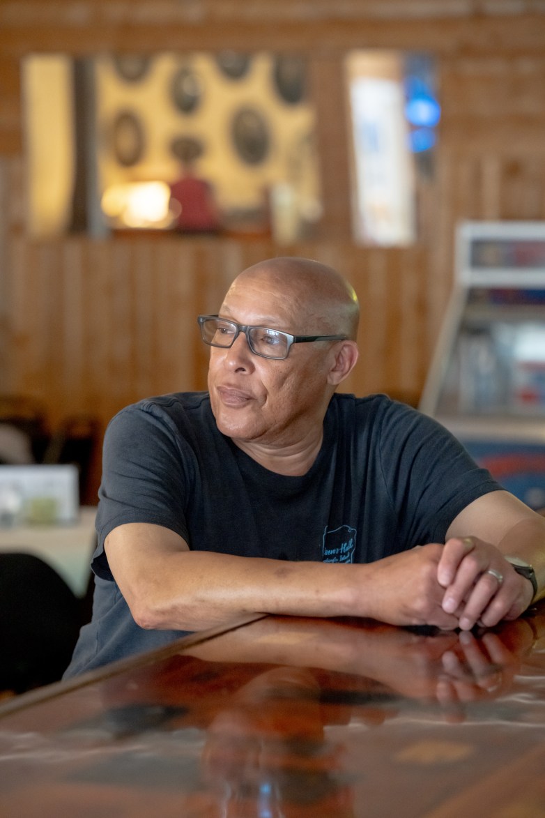 A person wearing glasses sits at a counter, looking to the side. Wood-paneled walls and seating are visible in the background.