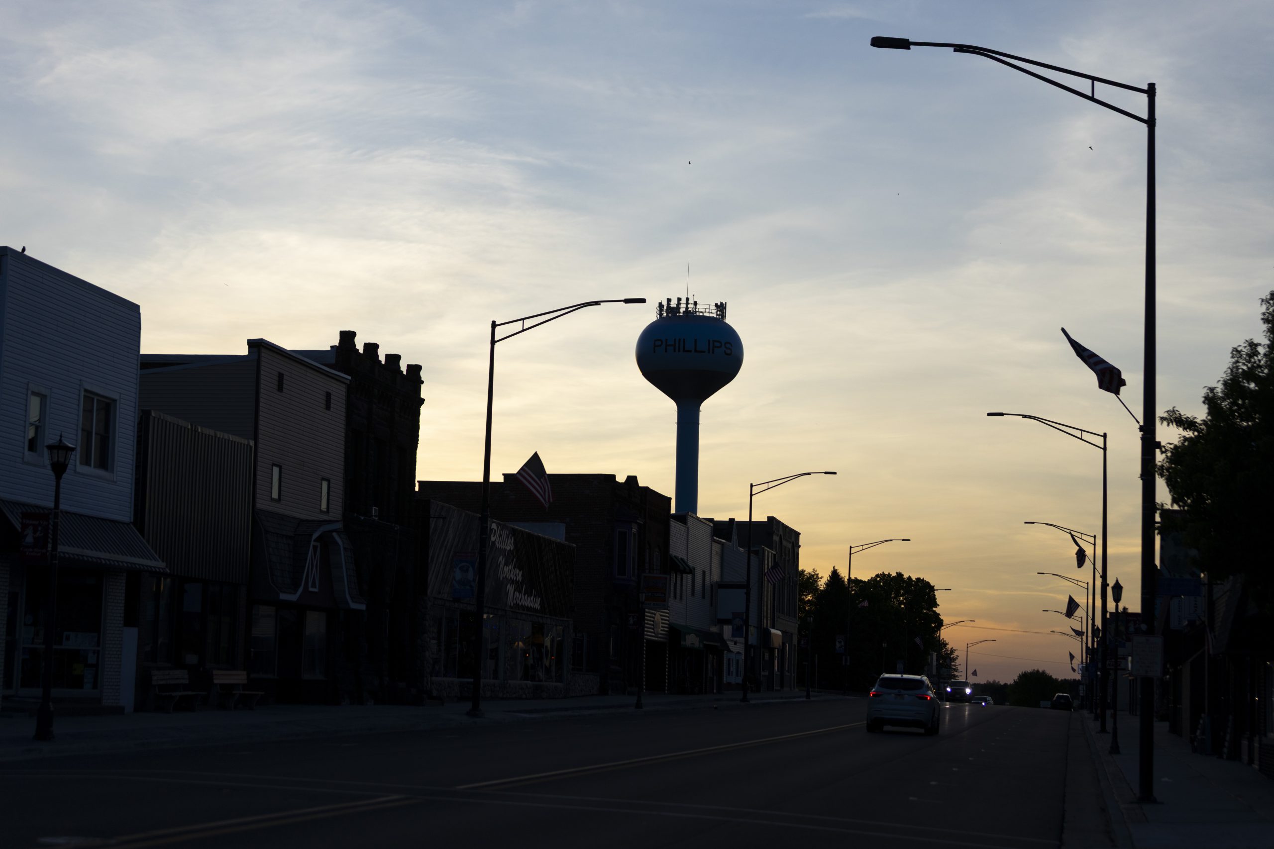 A water tower labeled “PHILLIPS” rises in silhouette above a downtown street lined with dark buildings, flags and streetlights at dusk.