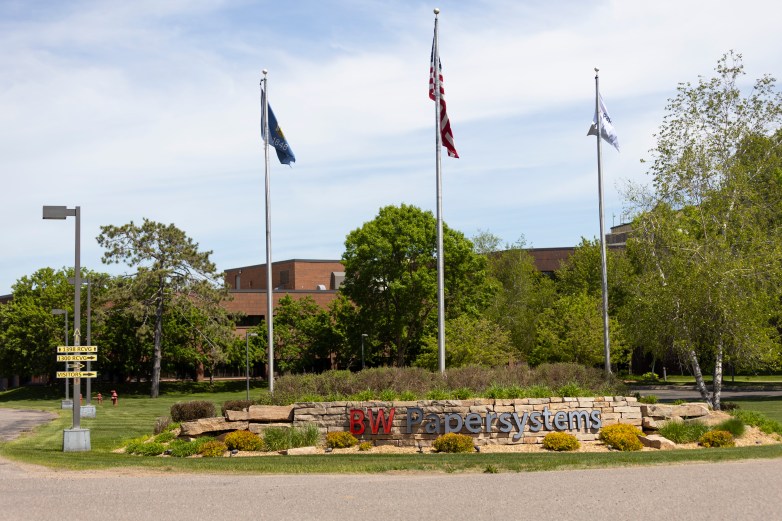 A stone sign reading “BW Papersystems” stands beside a roadway with three flagpoles, trees and office buildings in the background.