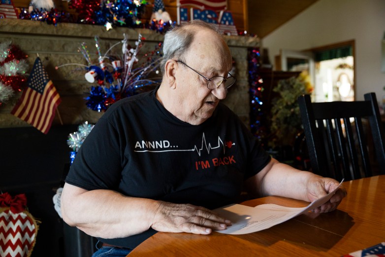 A person wearing a shirt reading “ANNNND... I’M BACK” sits at a table holding a document. Patriotic decorations and small American flags surround a fireplace in the background.