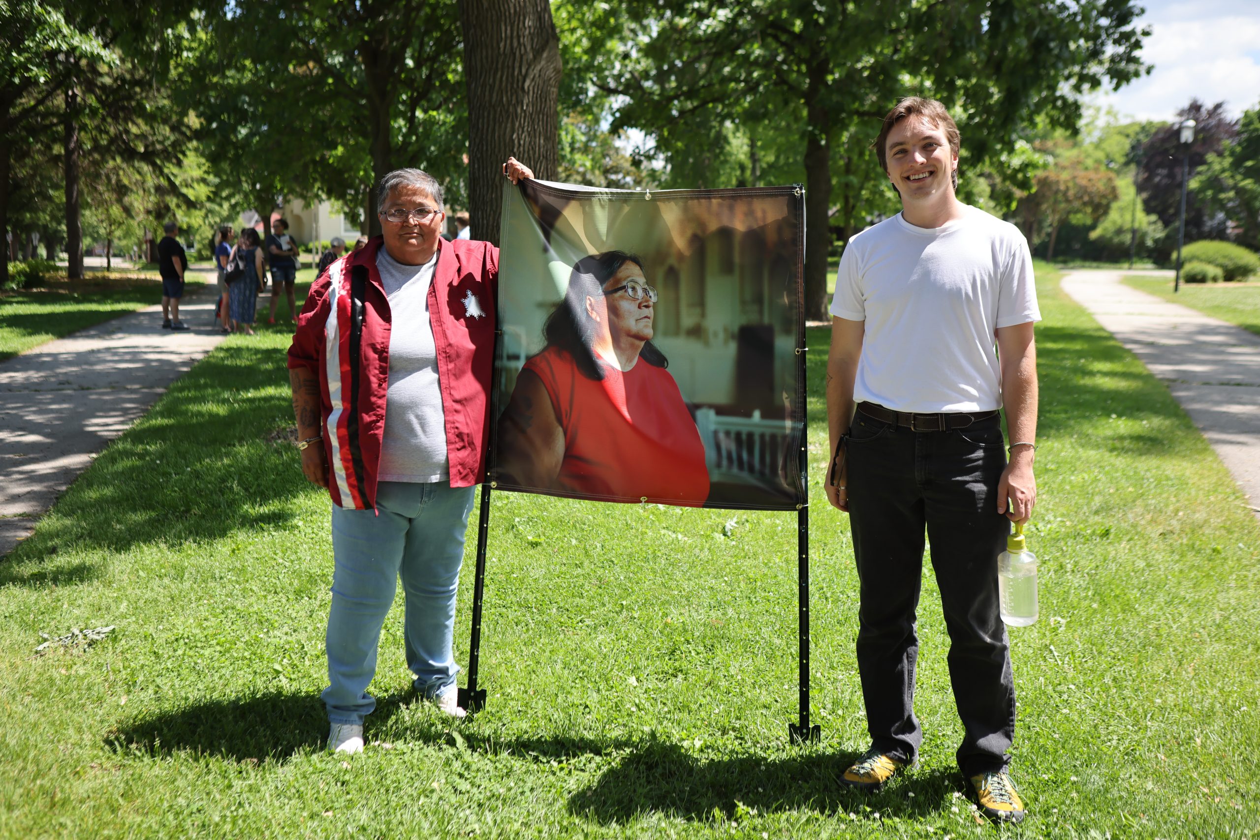Two people stand beside a freestanding display showing a portrait of a person in a red top, with trees, grass and a sidewalk in the background.