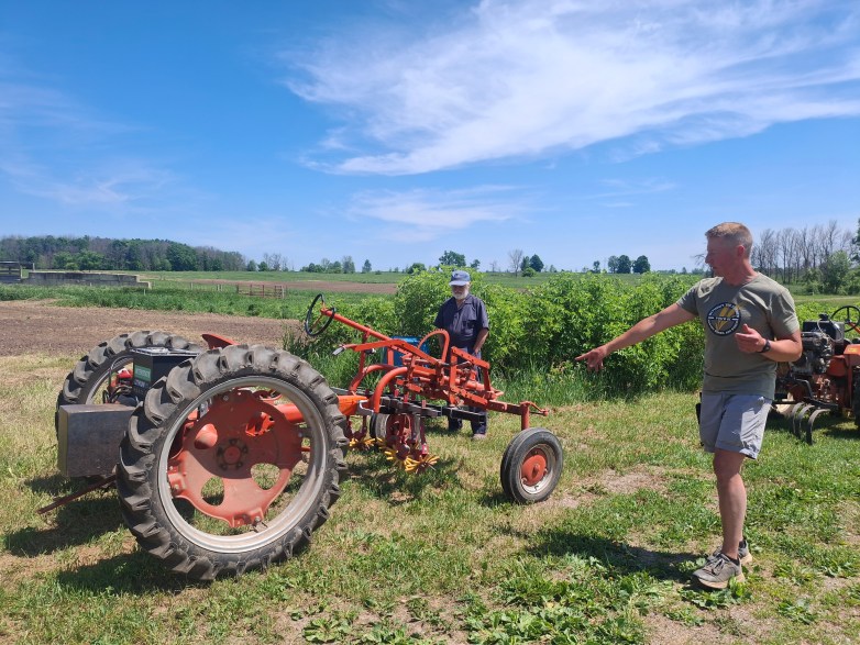 A person points toward a small red tractor in a field while another person stands beside the equipment, with rows of shrubs and open farmland behind them.
