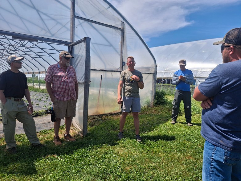 People stand on grass next to two greenhouses with a door open to one of the greenhouses under a blue sky.