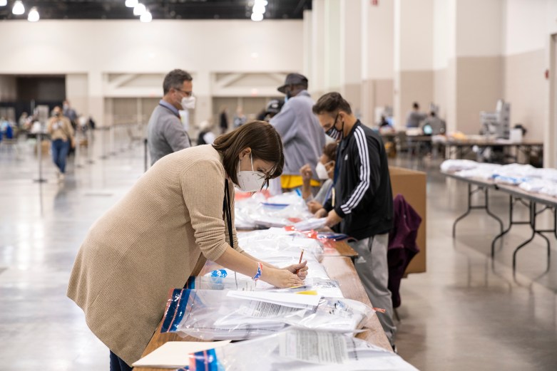 A person wearing a face mask writes on paperwork at a table covered with plastic bags while other people stand and sit in a large indoor hall