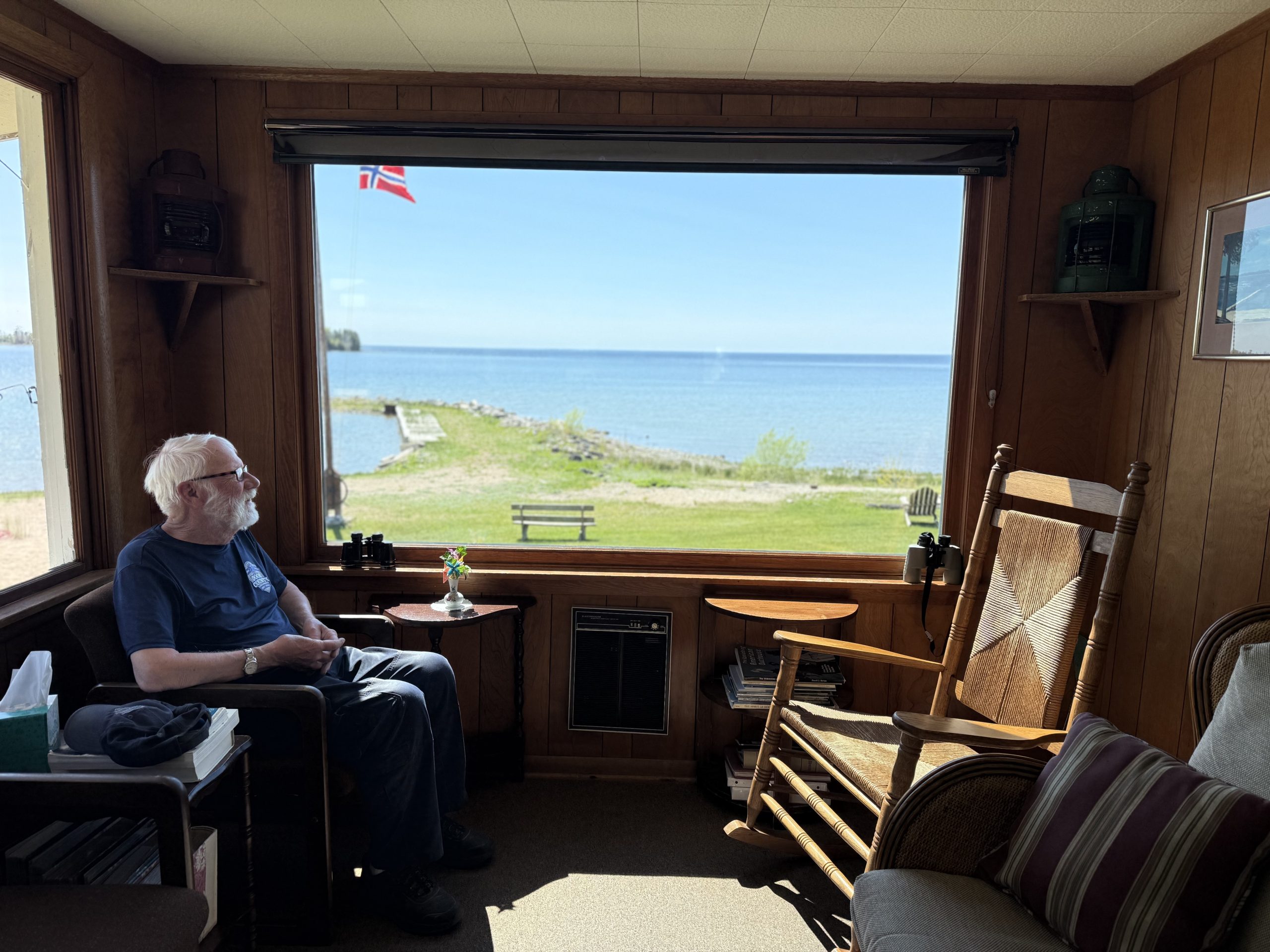 A person sits in a chair facing a large window overlooking a lakeshore, dock and open water. A rocking chair and small tables sit inside the wood-paneled room.