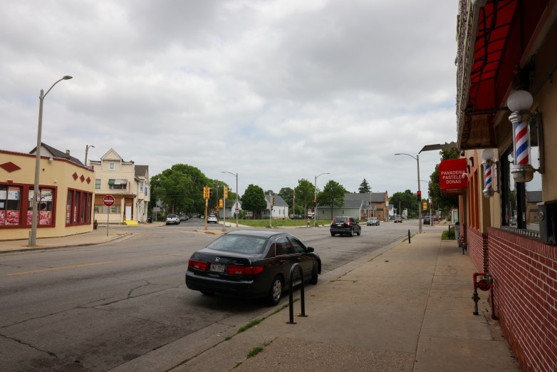 A street intersection with parked and moving cars is lined with storefronts and houses. A sign on one storefront reads 