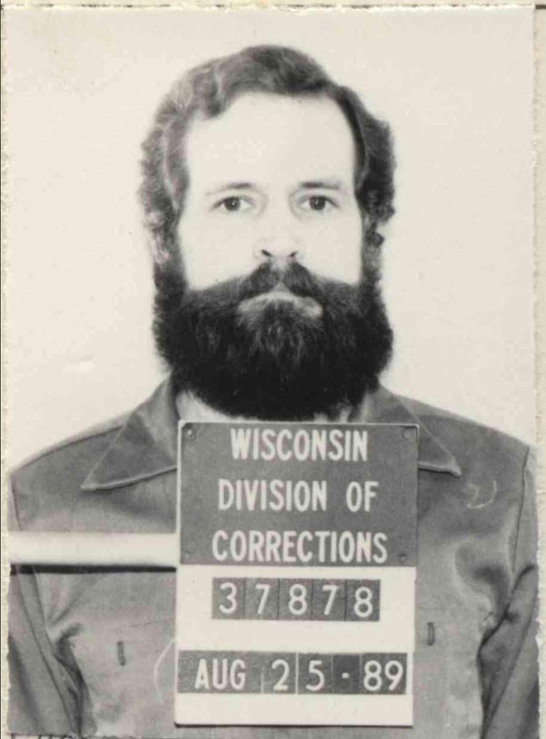 A person with a beard faces the camera behind a placard reading “WISCONSIN DIVISION OF CORRECTIONS” with identification number 37878 and the date “AUG 25-89.”