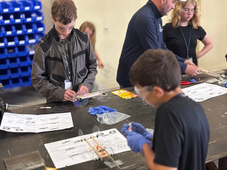 People assemble small wheeled model vehicles at a workbench with printed instructions.