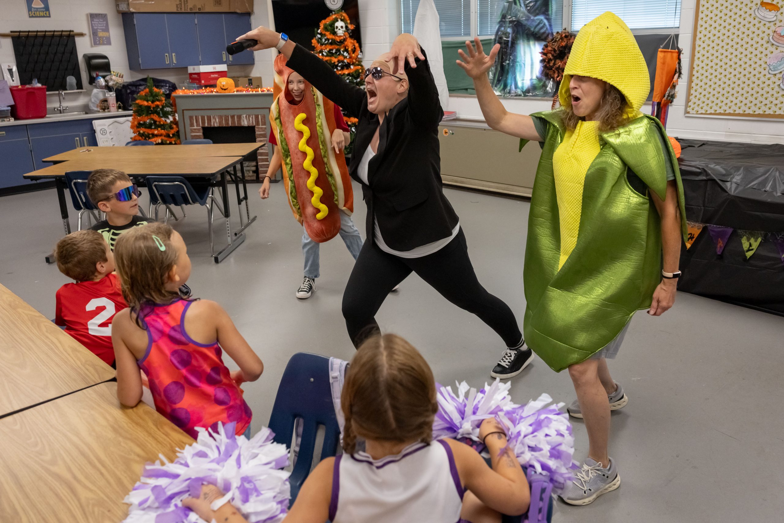 People in costume stand with raised arms in a classroom while children watch. Costumes include an ear of corn, a hot dog and a black outfit with sunglasses.