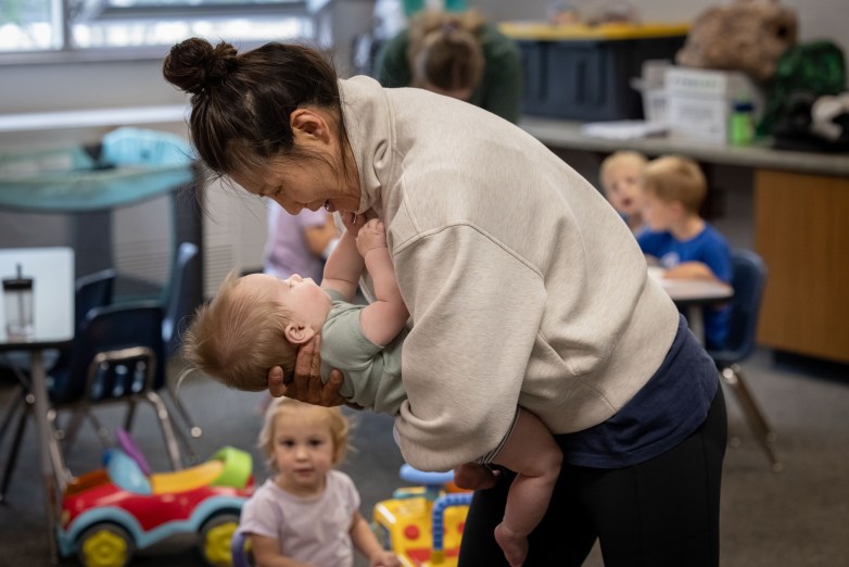 A person holds an infant and smiles while looking down at the child in a classroom. Other children sit in the background.