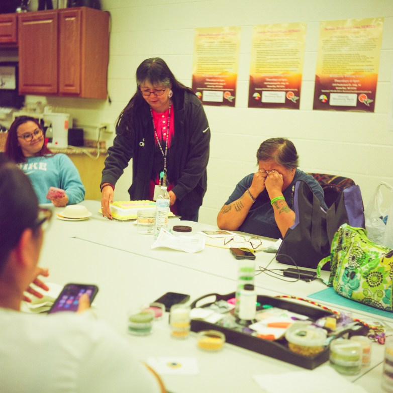 People sit around a table in a room while one person places a cake on the table. Another person sits with hands covering their face, and posters hang on the wall behind them.
