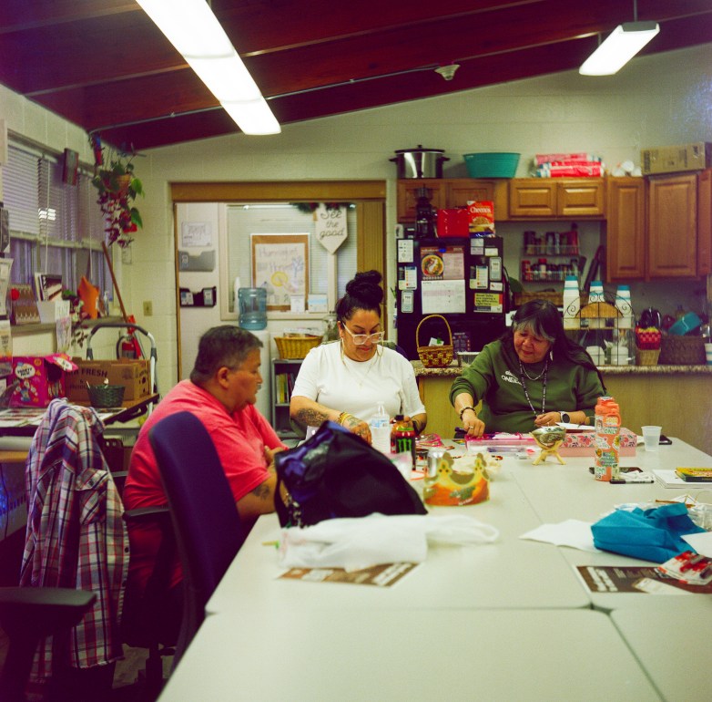 Three people sit around a table with craft supplies, drinks and personal items in a room with a kitchen area in the background.