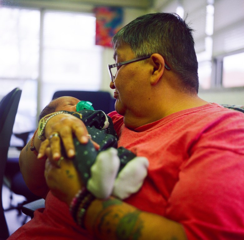A person wearing glasses and a red shirt holds a baby with a pacifier while seated indoors. Tattoos and bracelets are visible on the person's forearms.