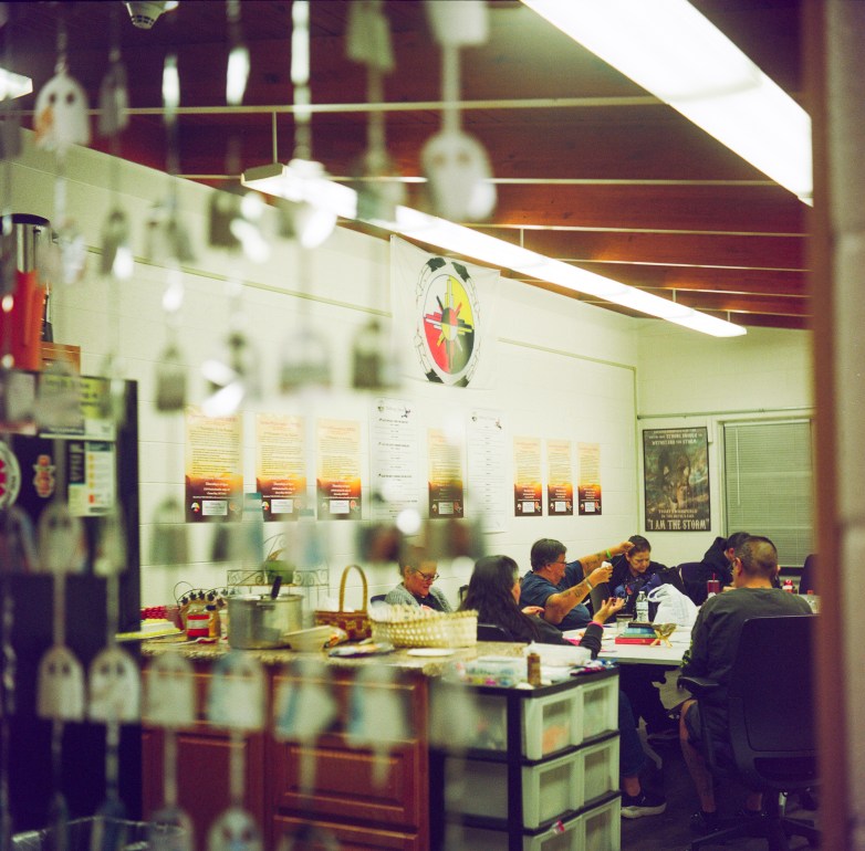 People sit around a table in a room with posters on the wall. Hanging decorations are out of focus in the foreground, partially framing the scene.