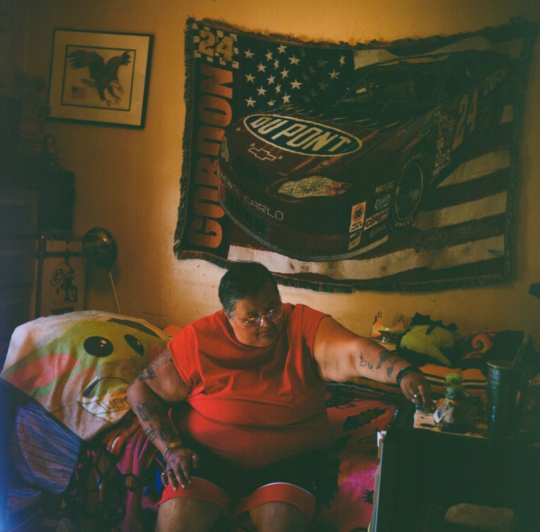 A person wearing a red shirt sits on a bed reaching toward a small table. A blanket with a race car and American flag hangs on the wall behind the person, with stuffed toys nearby.