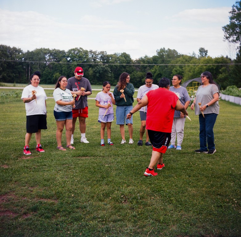 People stand in a line on a grassy field holding handheld items while one person in a red shirt walks toward them.