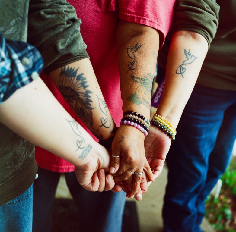 Four people stack their hands together. Matching bird-and-heart tattoos are visible on their forearms, along with bracelets and other tattoos.