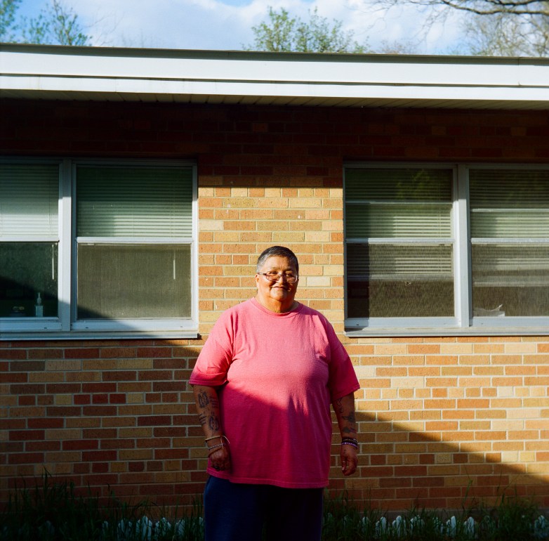 A person wearing a pink shirt stands in front of a brick building with two windows. Tattoos and bracelets are visible on the person's forearms.