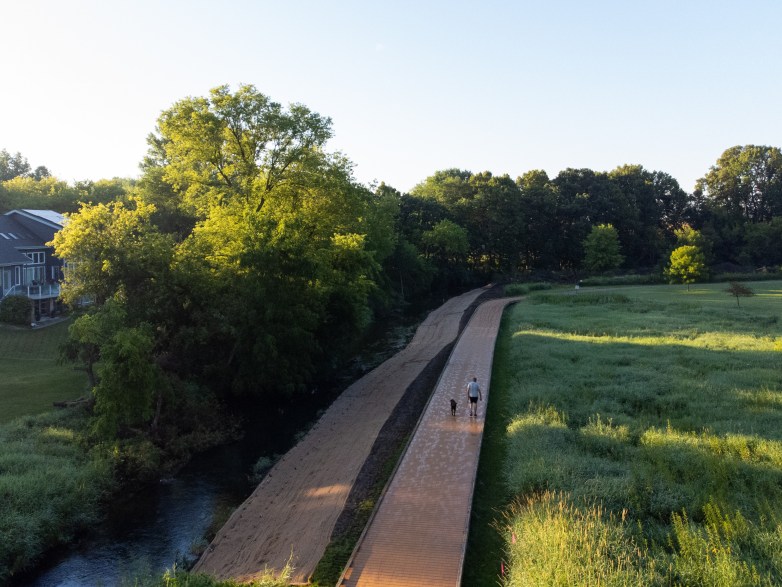 A person walks a dog on a paved path beside a narrow river, with grassy fields, trees and nearby houses under a clear sky.