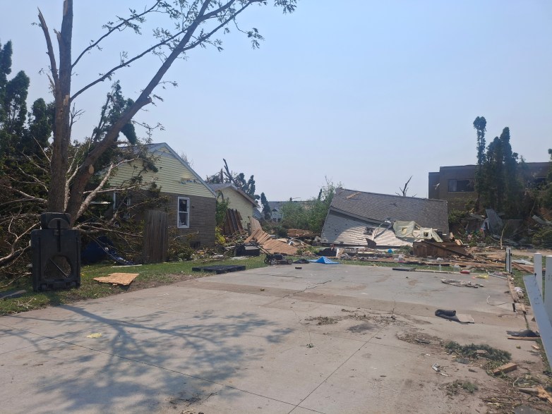 A damaged house and collapsed garage sit beside a driveway strewn with debris. Broken trees and scattered building materials fill the surrounding yard.