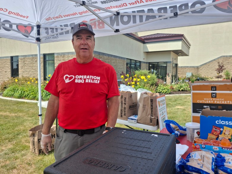A person wearing a shirt reading "Operation BBQ Relief" stands under a canopy beside food supplies, boxes, and a large cooler outside a building.