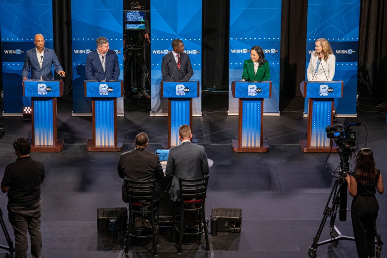 Five people stand at podiums on a stage with blue backdrops displaying the "WISN 12" logo. Camera operators and moderators are visible in the foreground.