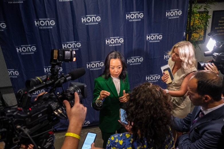 A person in a green suit speaks with people holding microphones and smartphones in front of a backdrop reading "FRANCESCA HONG FOR GOVERNOR."