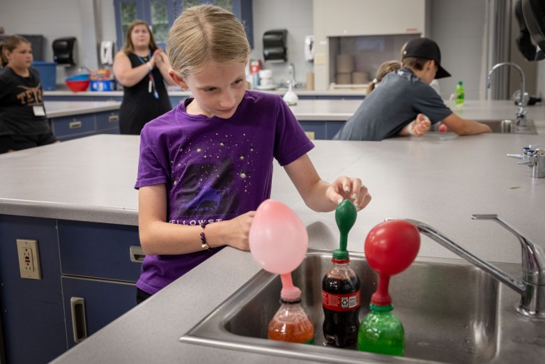 A person hovers one hand over a balloon at the top of a plastic bottle beside other bottles with inflated balloons in a sink. Other people are in the background.