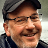 A person wearing glasses, a dark cap and a dark jacket smiles near a body of water with trees along the opposite shore.
