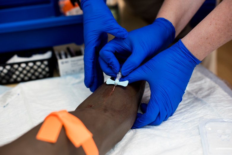 Blue-gloved hands insert a needle into a training arm with an orange tourniquet on a table covered with medical supplies.
